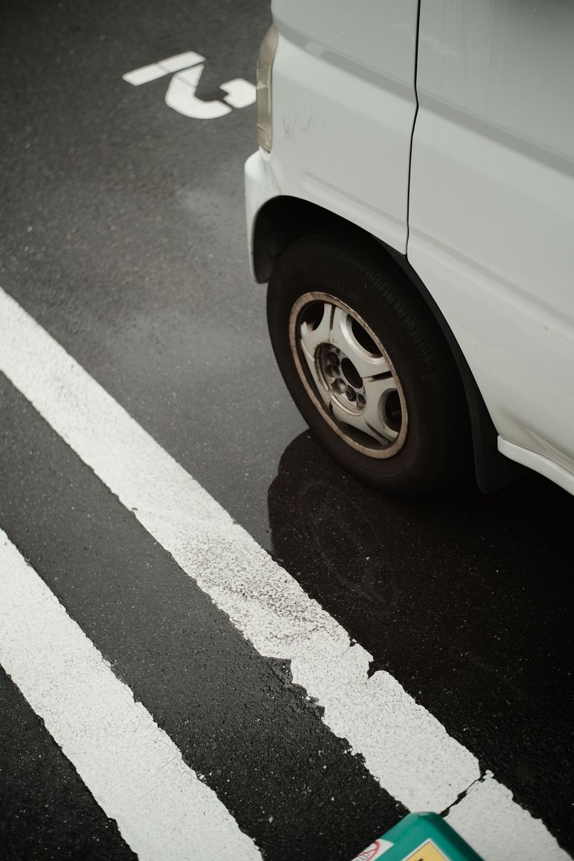Uma van branca estacionada em uma vaga demarcada e molhada, com uma linha branca tracejada visível no asfalto.