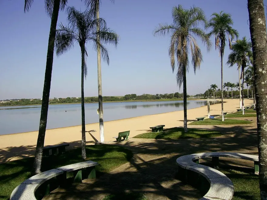 Praia ensolarada com palmeiras, guarda-sóis e água azul calma sob um céu parcialmente nublado.