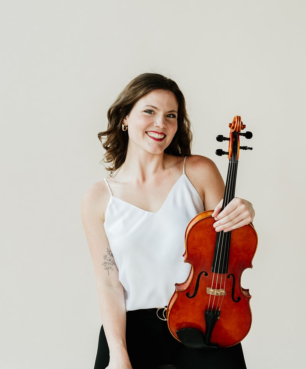 A woman in a white tank top is holding a violin and smiling.