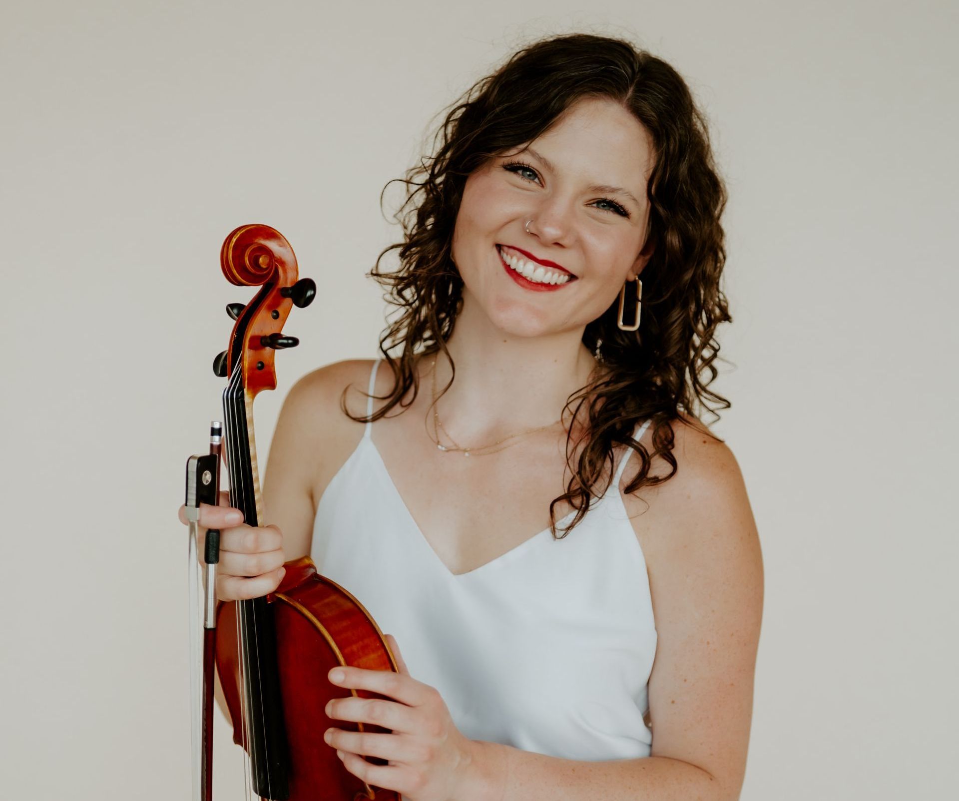 A woman in a white tank top is holding a violin and smiling.