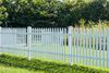A white picket fence stands in front of a green lawn and trees on a sunny day.