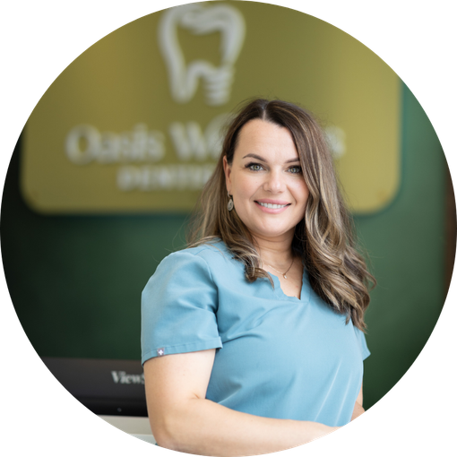 Dr Shuba at Oasis Wellness Dentistry, in a blue dental uniform poses in front of a dental office sign.