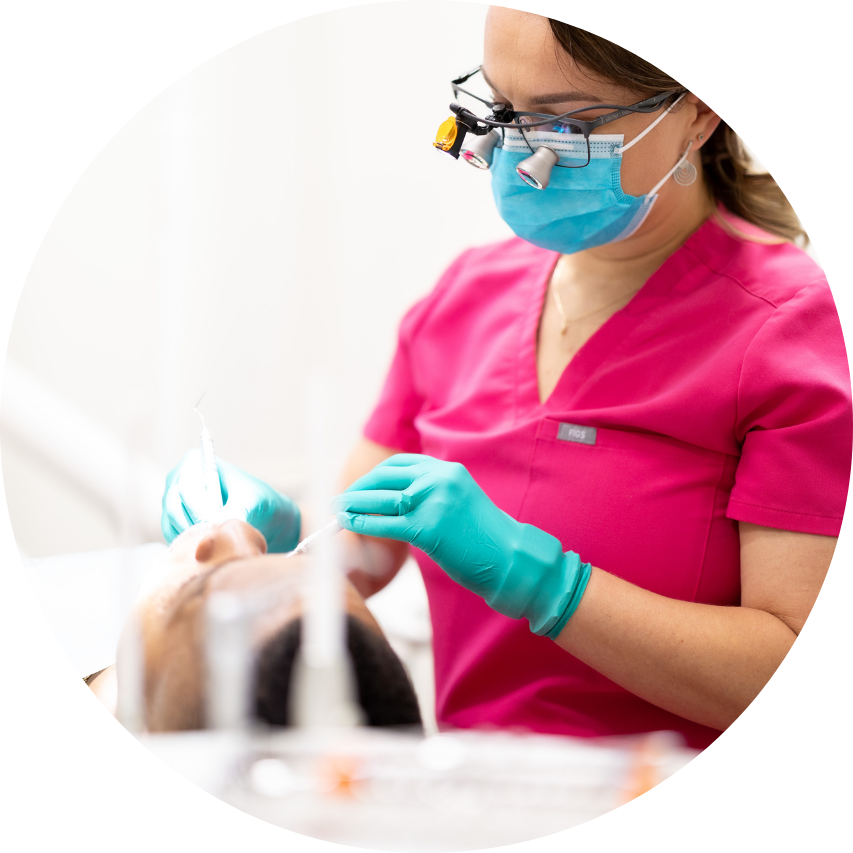 Dentist in pink scrubs, mask, and loupes examining a patient's mouth.