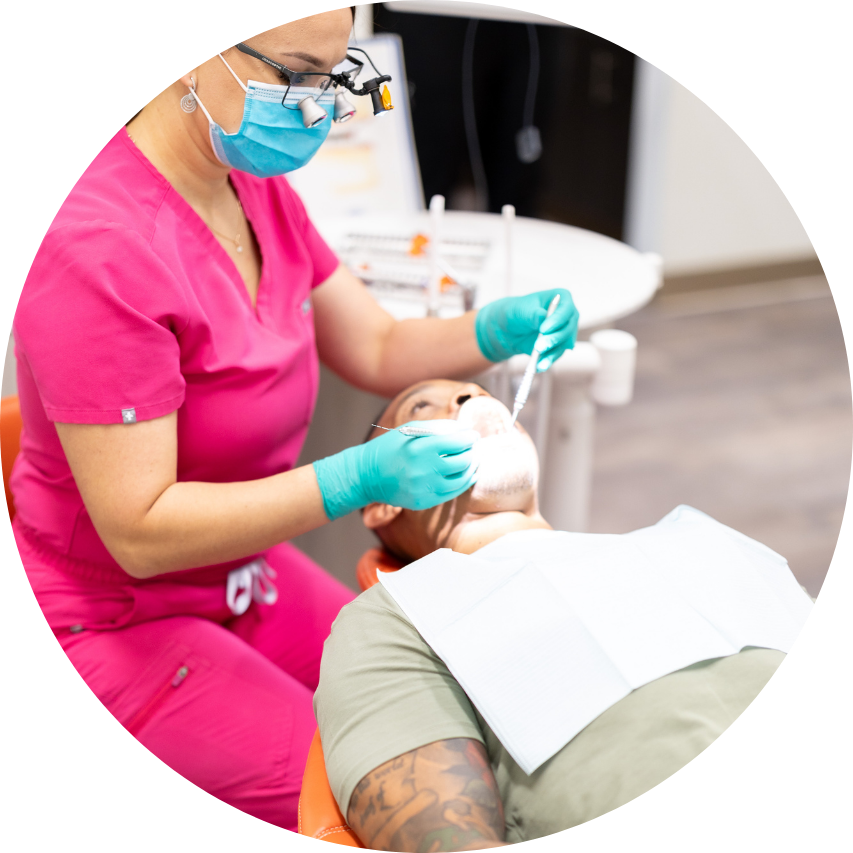 Dentist in pink scrubs, mask, and loupes examining a patient's teeth.