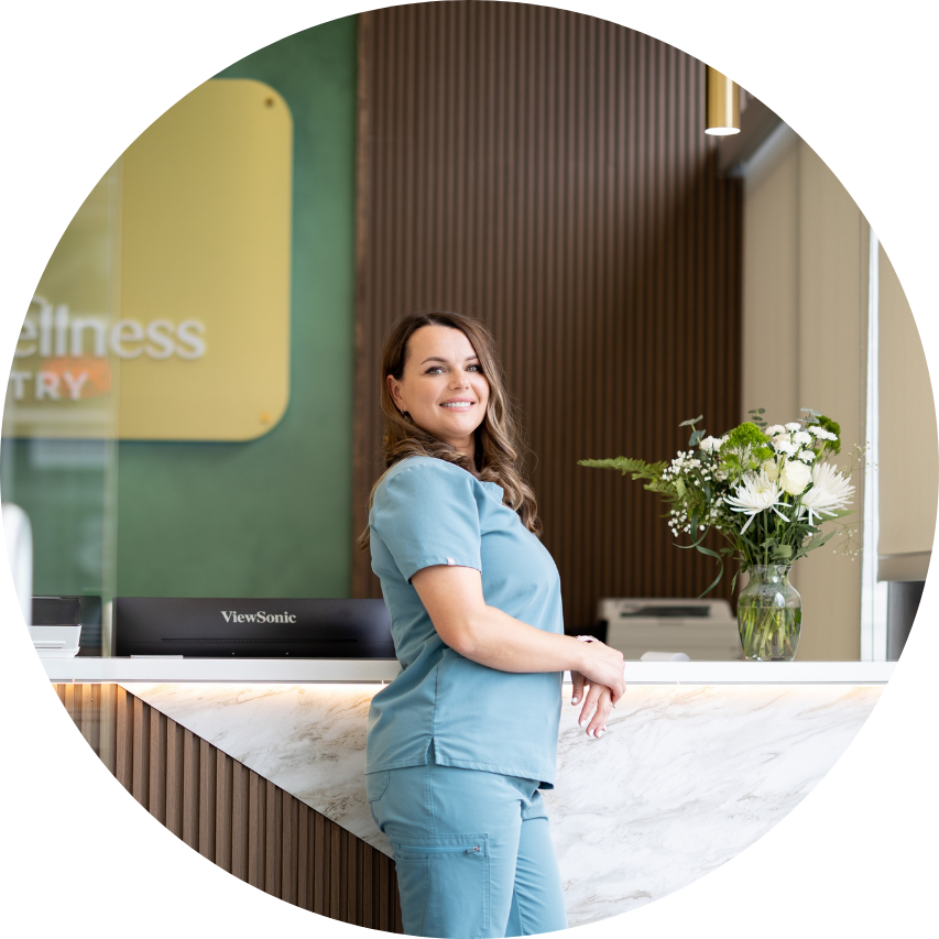 Woman in blue scrubs smiles near a reception desk in a modern dental office.