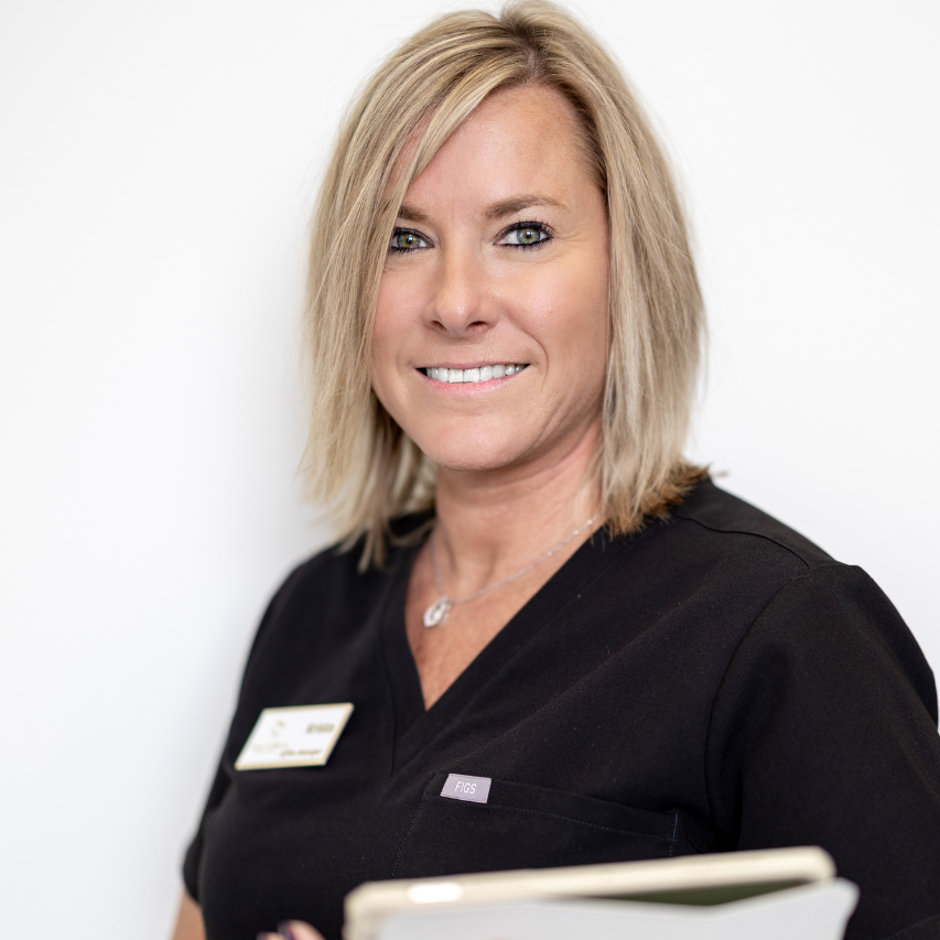 Woman with blonde hair wearing a black scrub top, smiling, in a medical setting, holding papers.