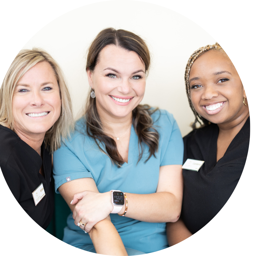 Three smiling medical professionals in front of a white background. One in a blue scrub, two in black scrubs.