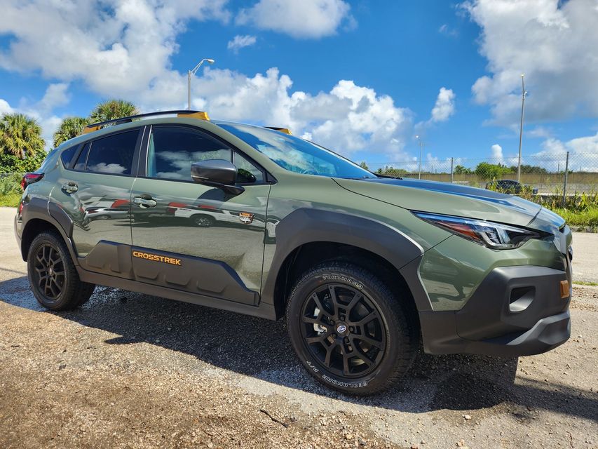 Green SUV with black wheels, roof rack, and side lettering parked on pavement under a cloudy sky.