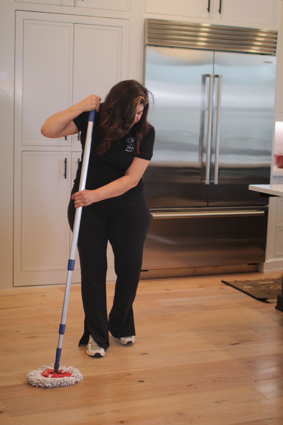 Woman mopping light wood floor in a kitchen with a stainless steel refrigerator and white cabinets. Woman mopping light wood floor in a kitchen with a stainless steel refrigerator and white cabinets.