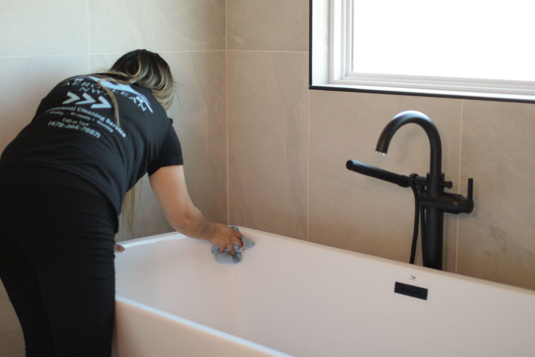 Person cleaning a white bathtub in a bathroom with beige tiles and black faucet. Person cleaning a white bathtub in a bathroom with beige tiles and black faucet.