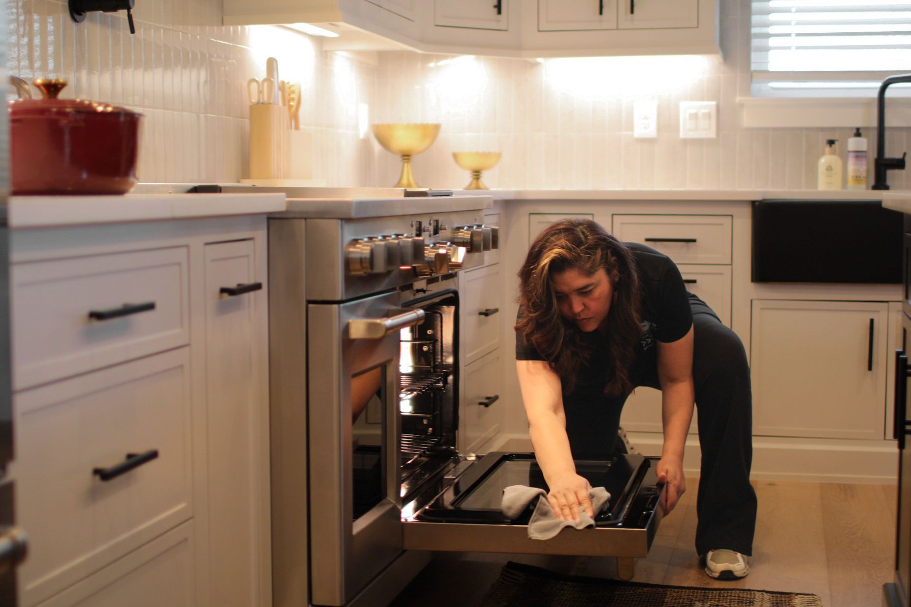 Woman cleaning the inside of a stainless steel oven in a white kitchen with black hardware. Woman cleaning the inside of a stainless steel oven in a white kitchen with black hardware.