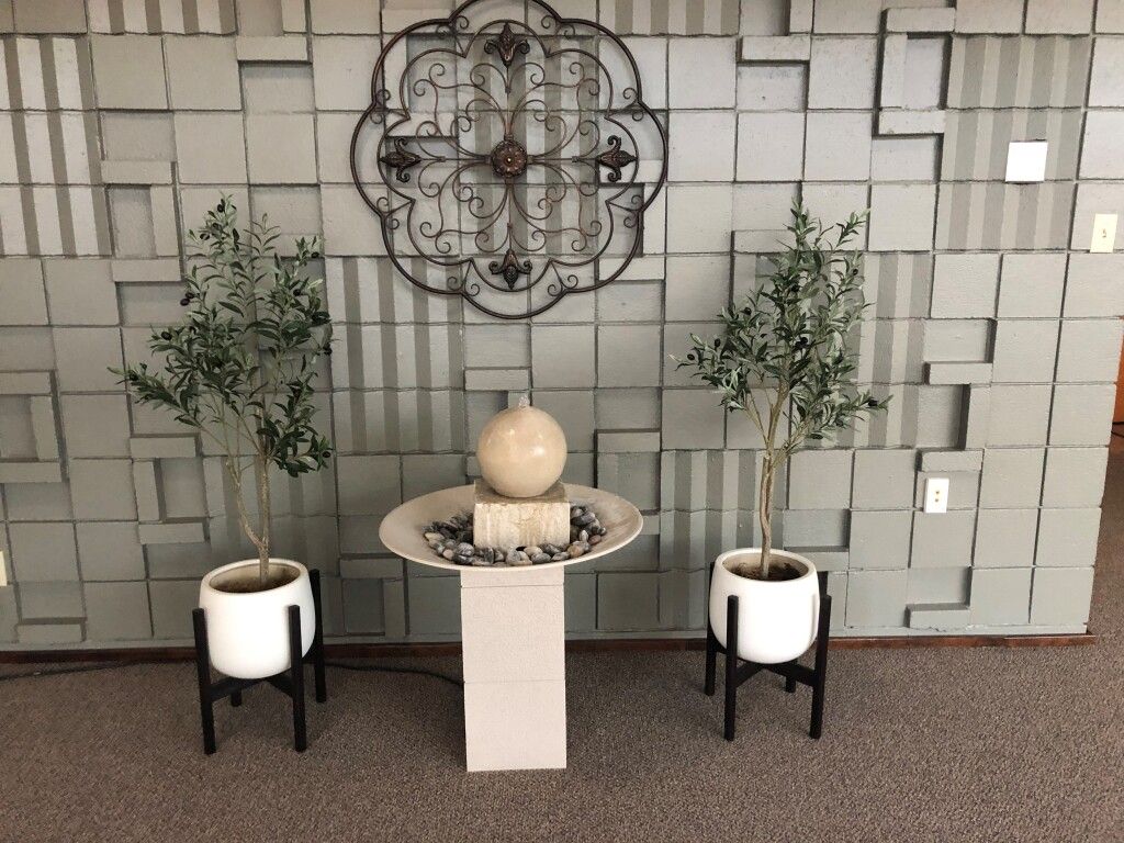 Two potted trees flank a fountain, all before a textured wall with ornate metal decor.