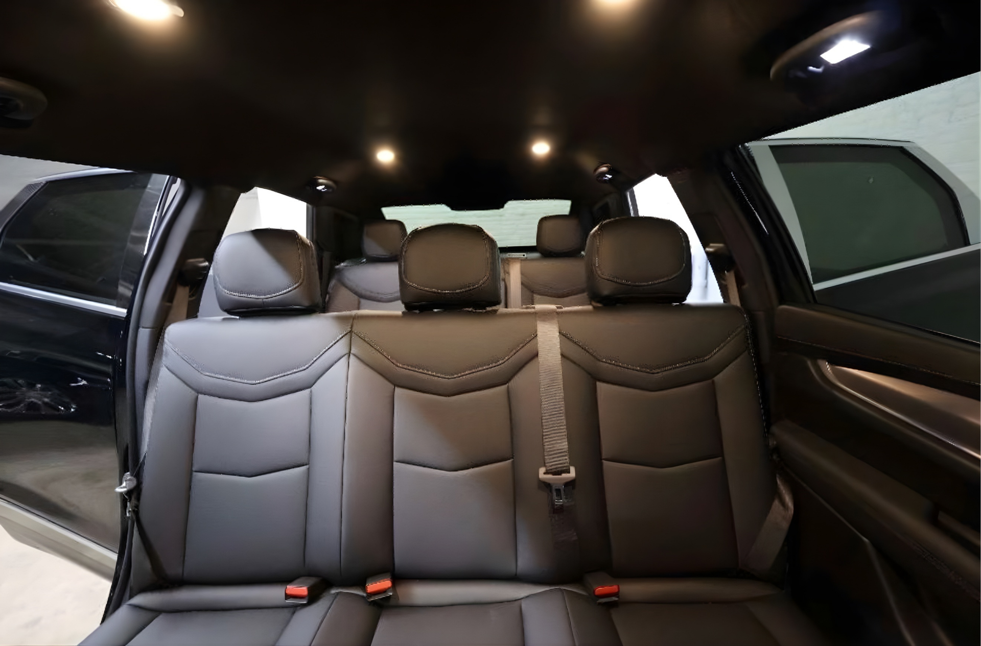 Interior view of a car's back seats: black leather, three seats, overhead lights, open doors.
