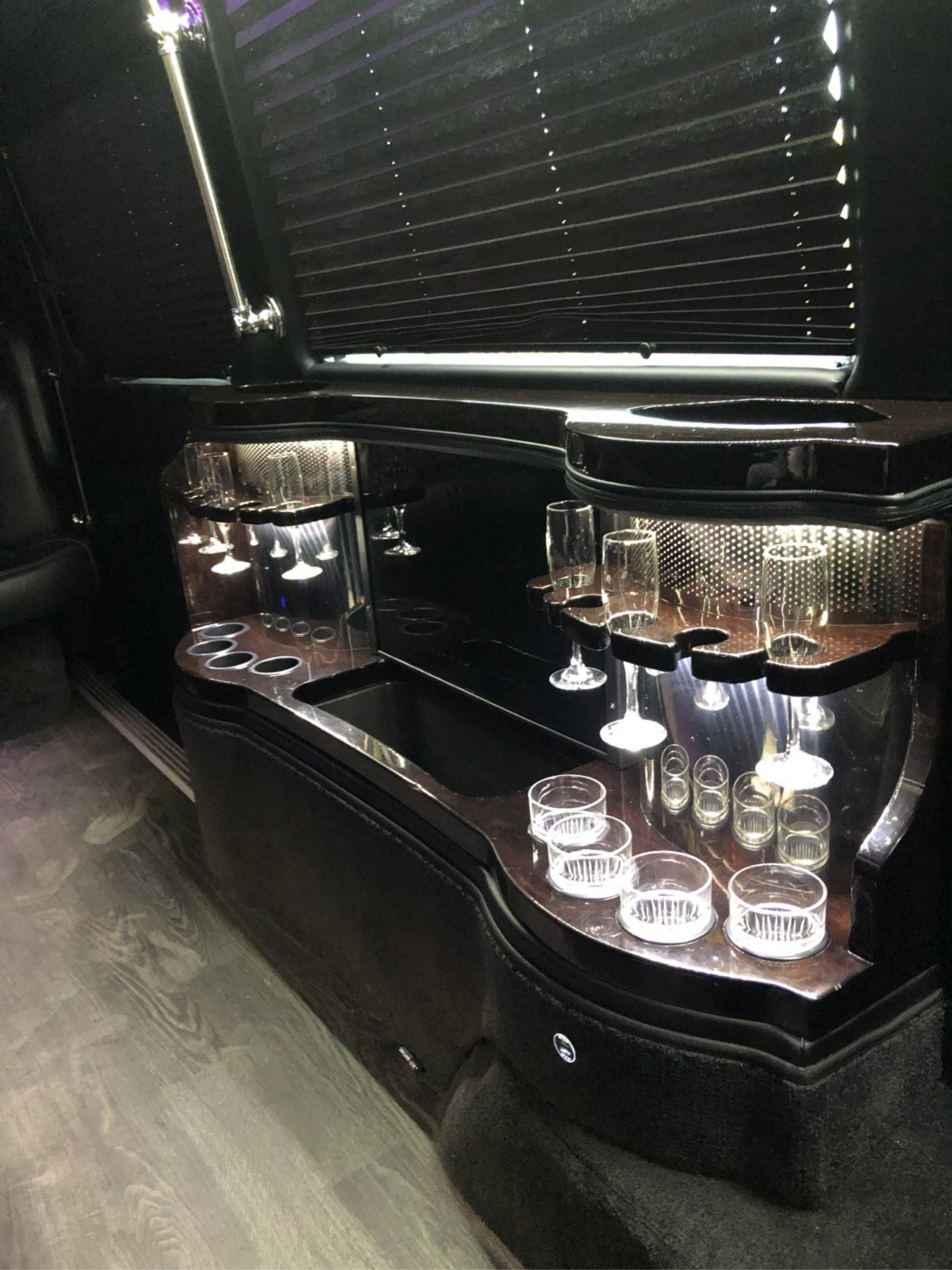 Interior of a limousine. Dark wood bar area with glasses, lighting, and a window blind.