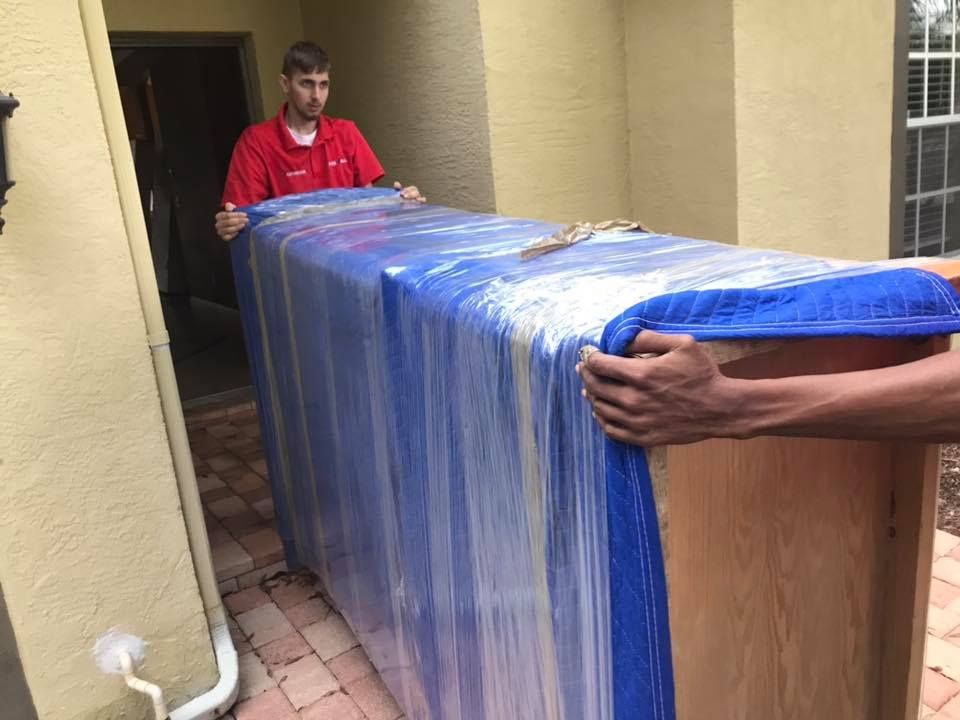 Two men are carrying a large piece of furniture wrapped in plastic.