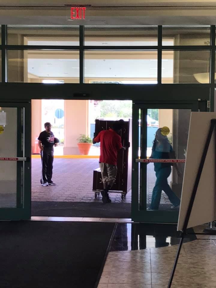 A man in a red shirt is pushing a piano through a glass door. 28.4845° N, 81.2519° W
