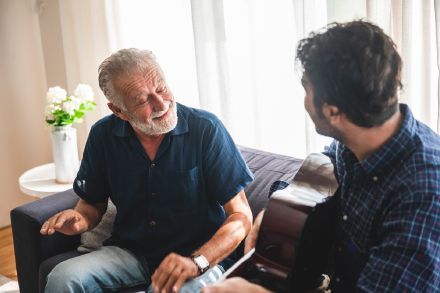 An older man is sitting on a couch talking to a younger man who is playing a guitar.