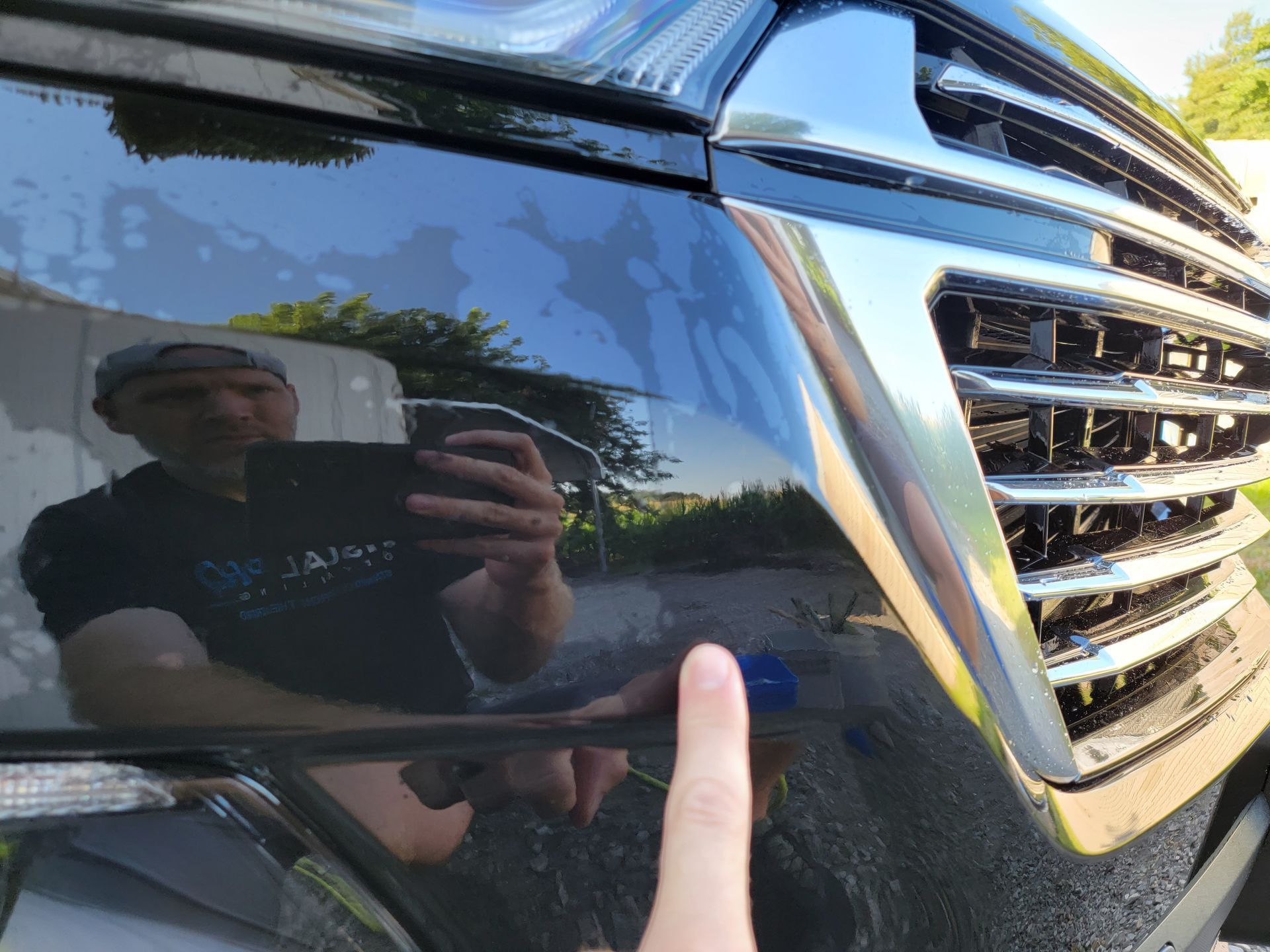 Close-up of a black car’s glossy front corner with chrome grille and a finger pointing at the paint finish