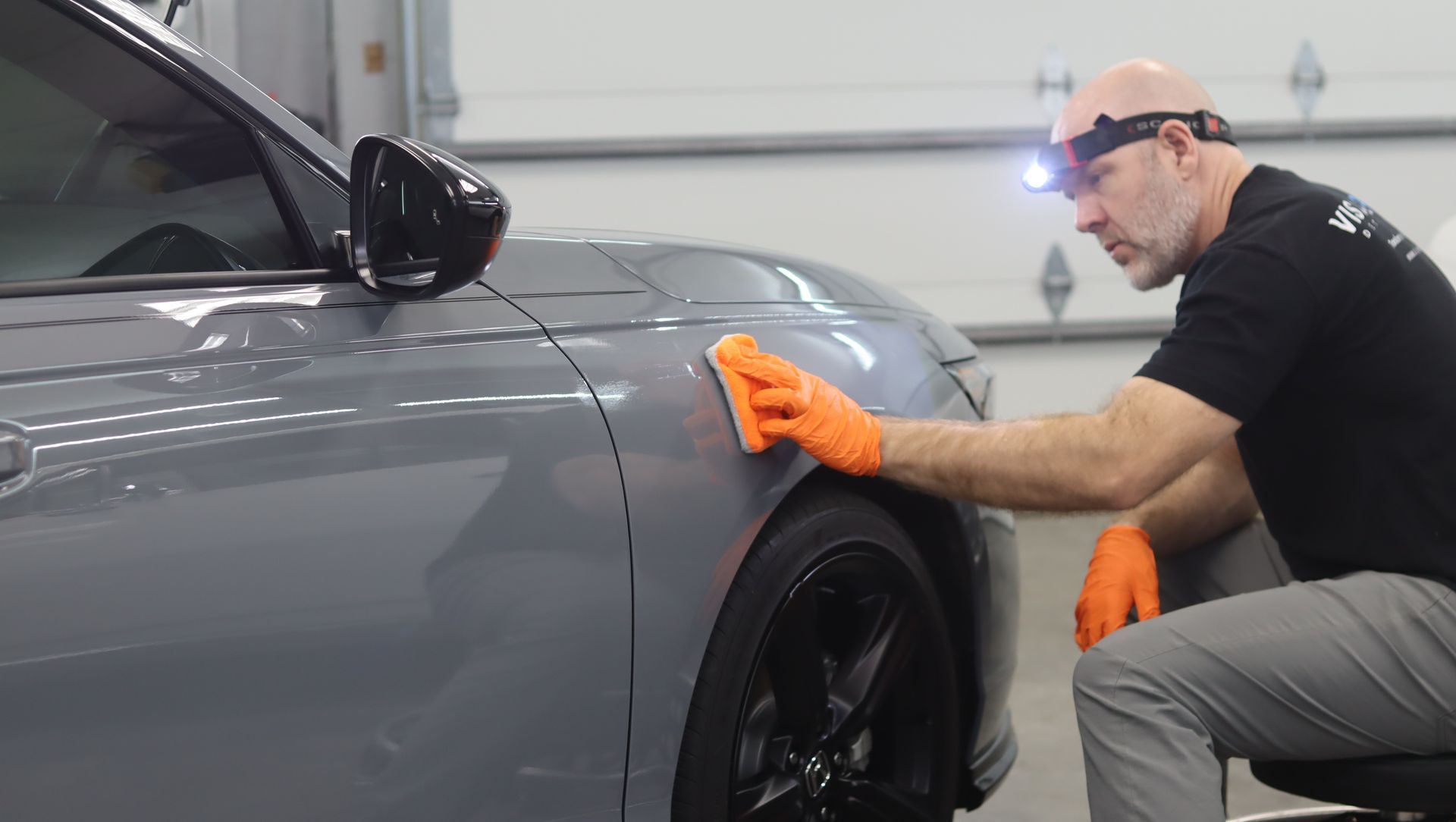 Technician in orange gloves polishing the fender of a gray car in a garage