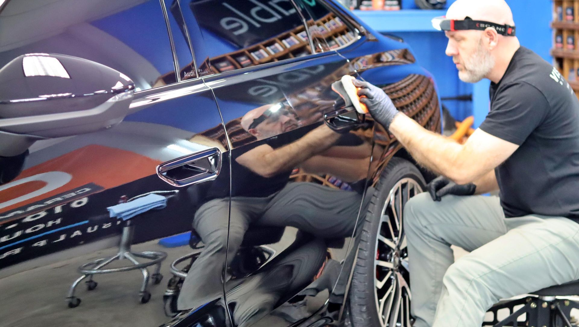 Man polishing a glossy black car in a workshop, reflected in the paint, with the wheel visible.