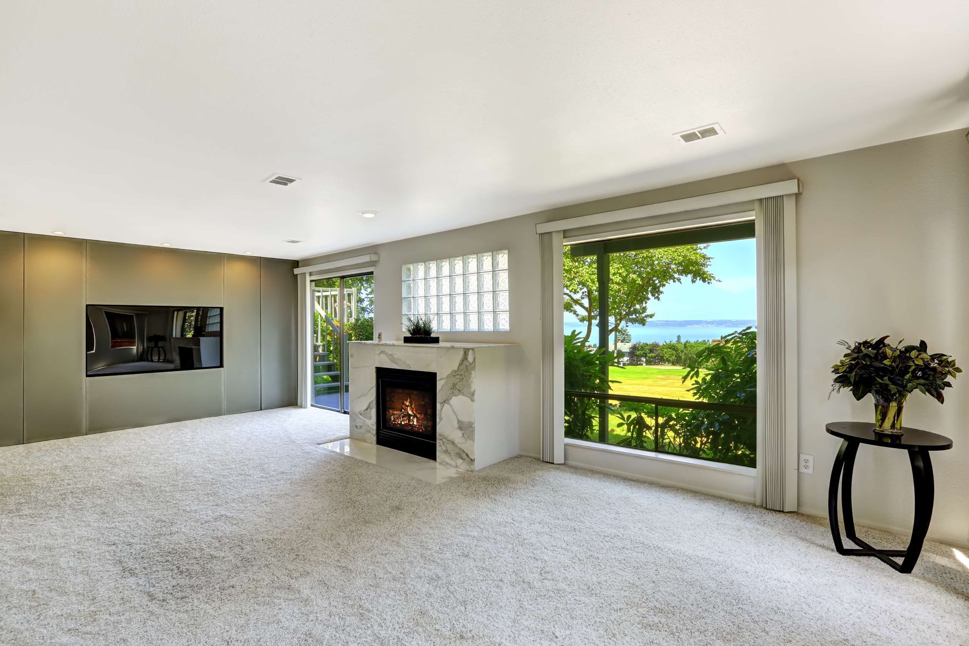 Bright living room with a fireplace, carpeted floor, and large windows overlooking green hills and ocean.