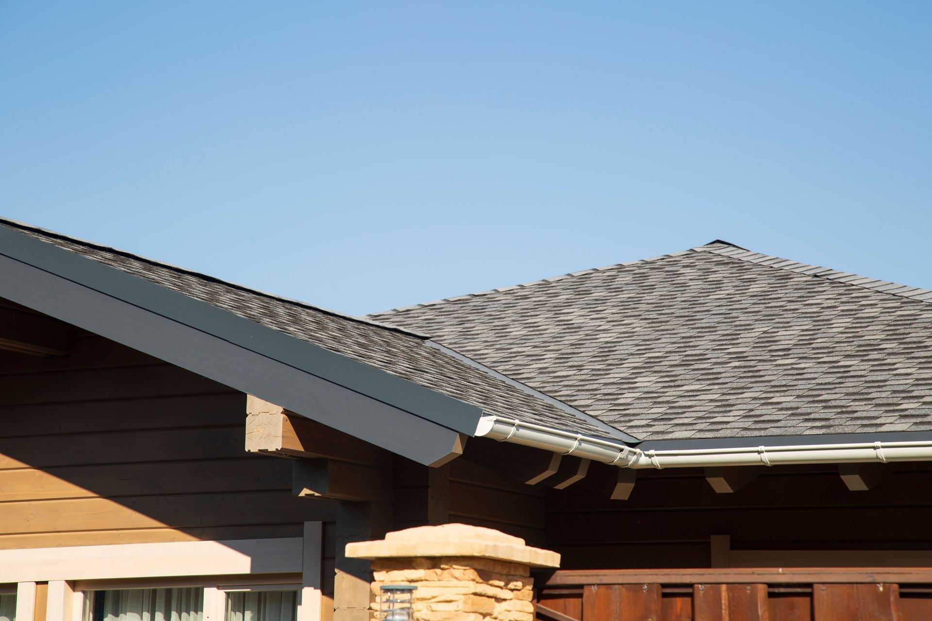 Roofline of a suburban house with gray shingles and tan siding against a clear blue sky