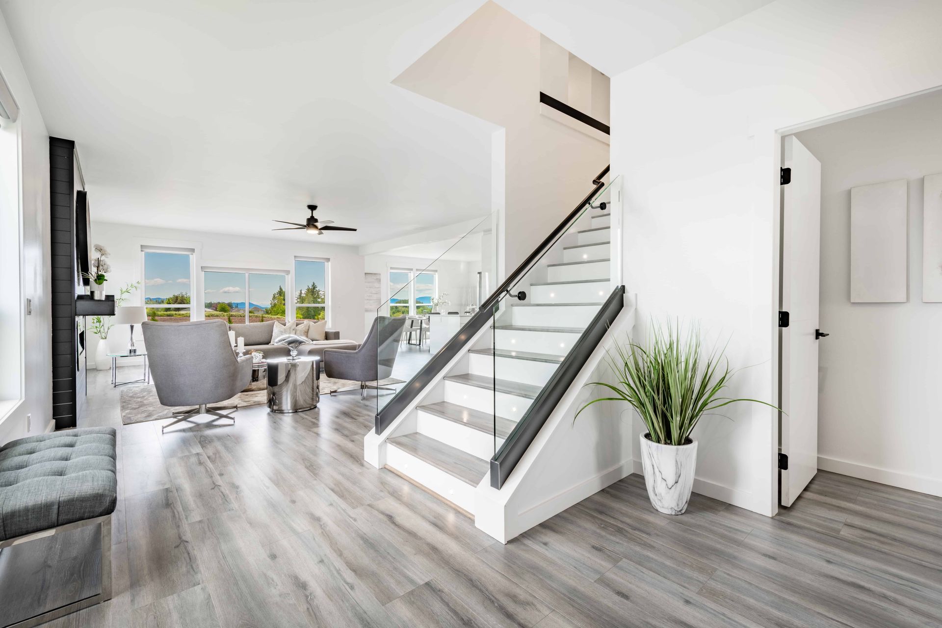 Bright modern living room with staircase, gray furniture, and large windows overlooking a city view