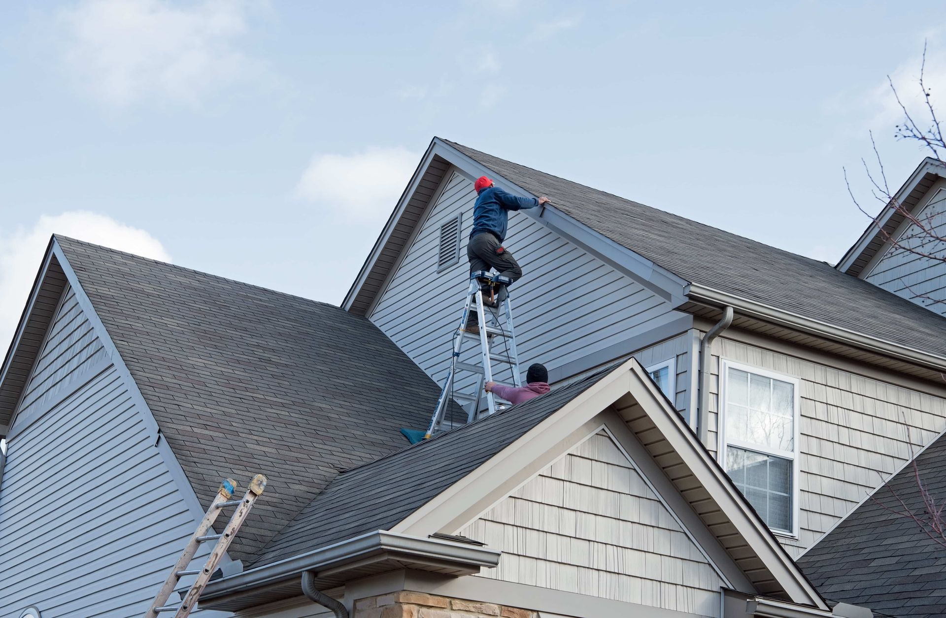 Two workers repairing a gray shingled roof on a house with a ladder.