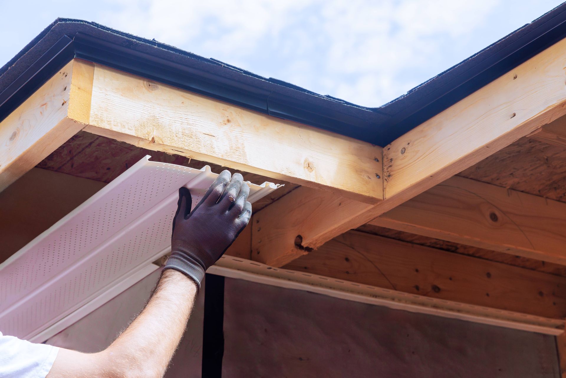 Gloved hand installing soffit under a house eave with exposed wooden framing