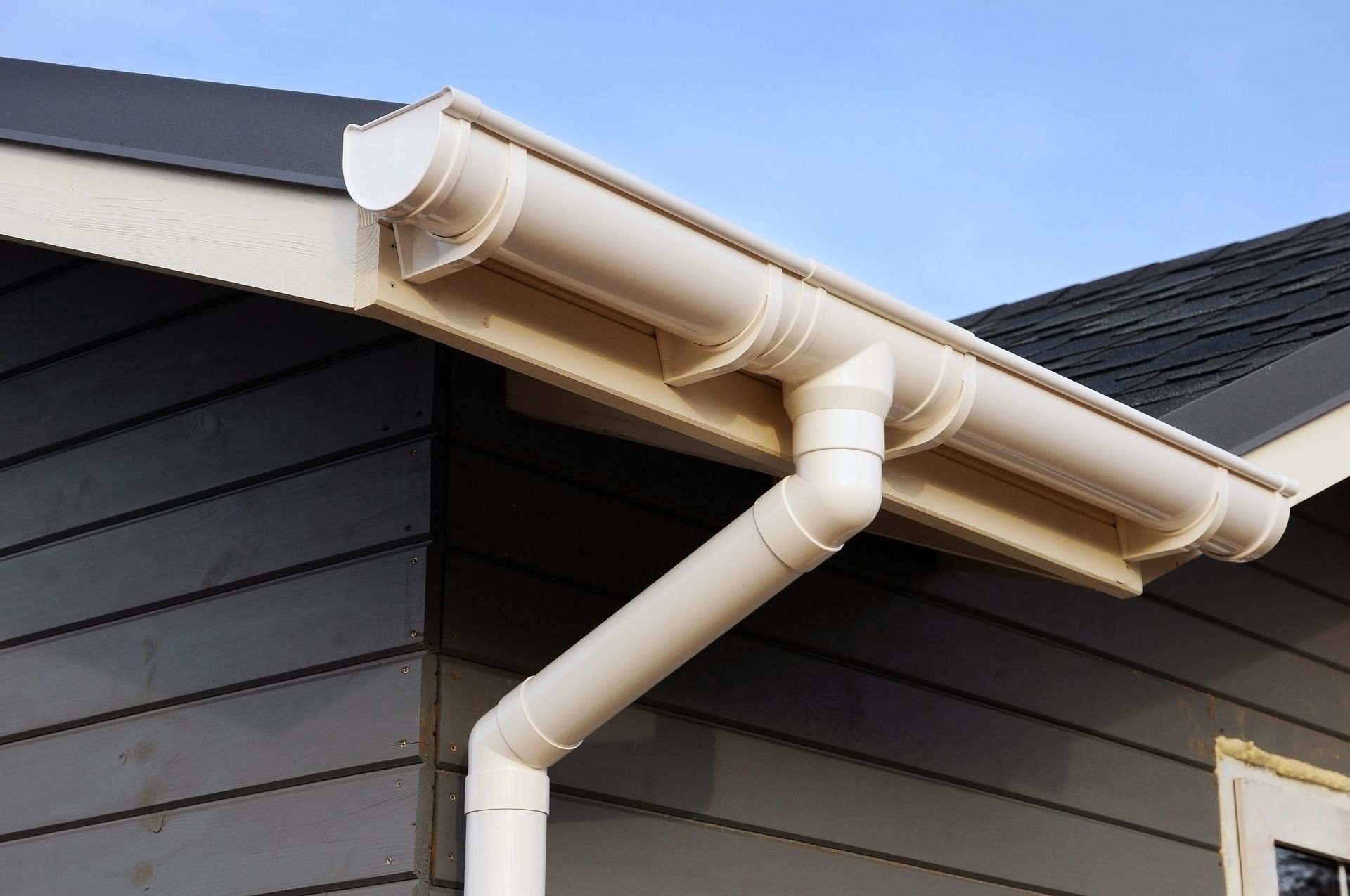 White rain gutter and downspout on a dark house exterior under a blue sky