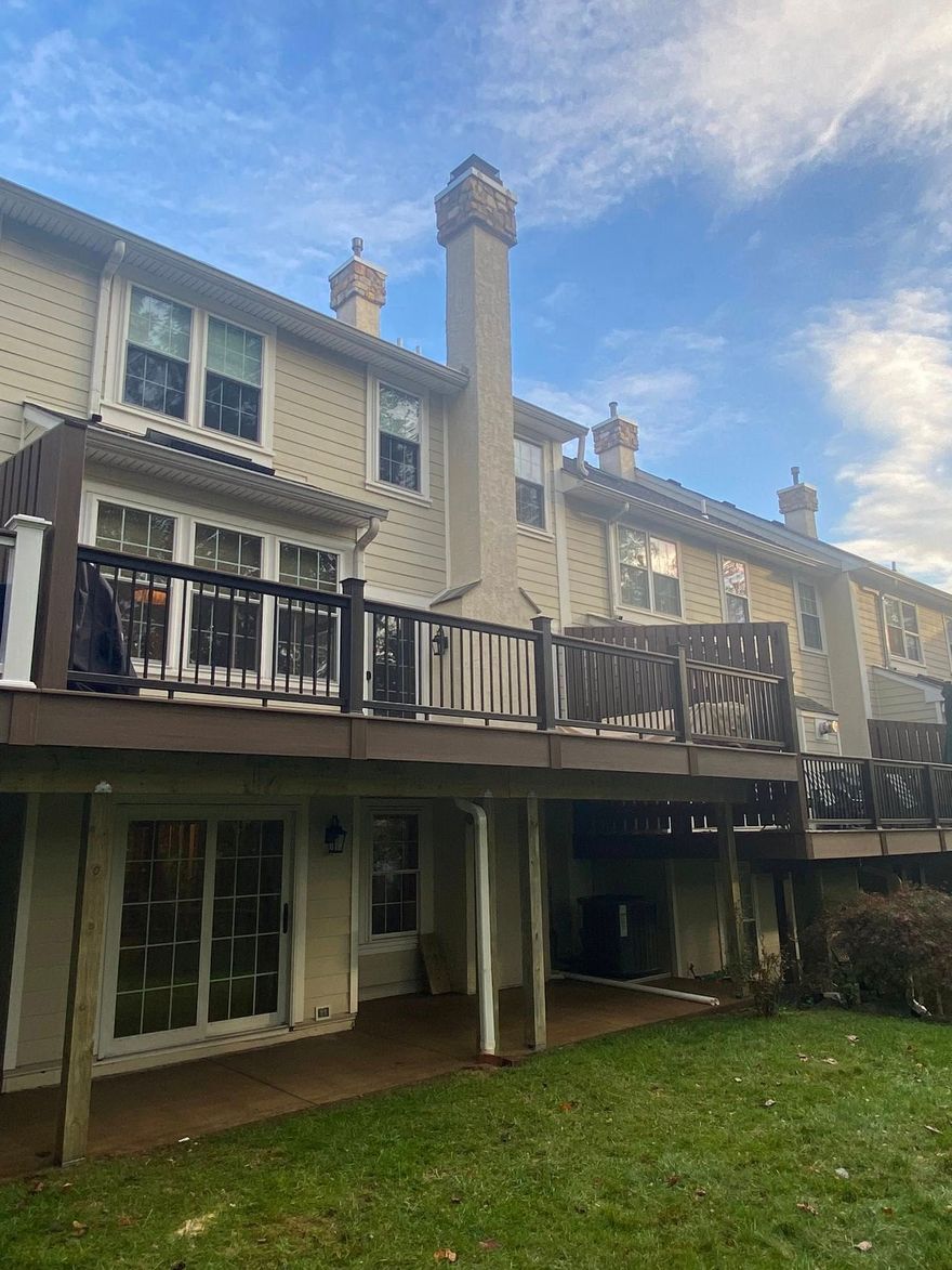 Back view of a beige townhouse with deck, sliding doors, chimneys, and a grassy yard under a cloudy sky