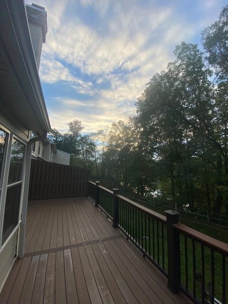 Wooden deck with dark railing beside a house, overlooking trees under a cloudy sunset sky.