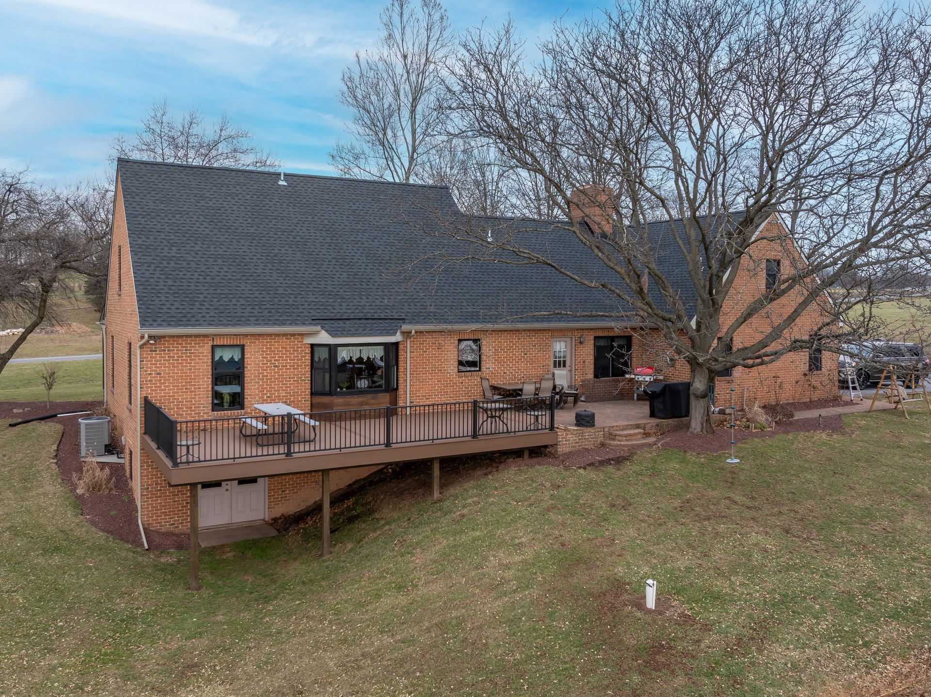 Brick house with dark roof and wooden deck on a grassy hill, surrounded by bare trees