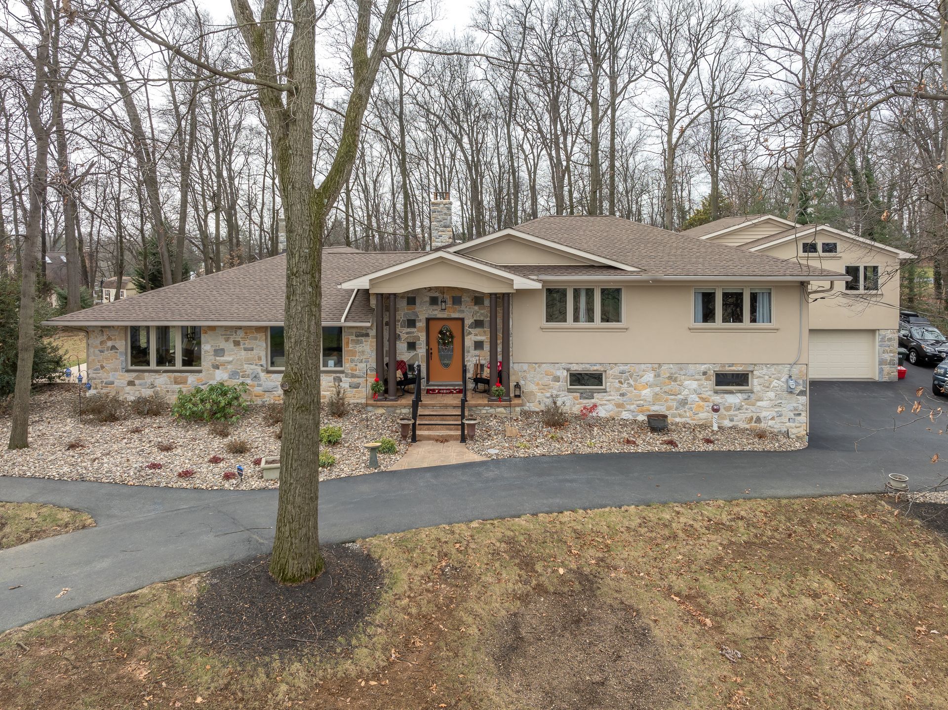 Single-story beige house with stone facade, front porch, and attached garage in a wooded yard