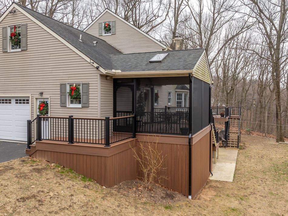 Brown house with a raised wooden deck and screened porch beside a wooded yard