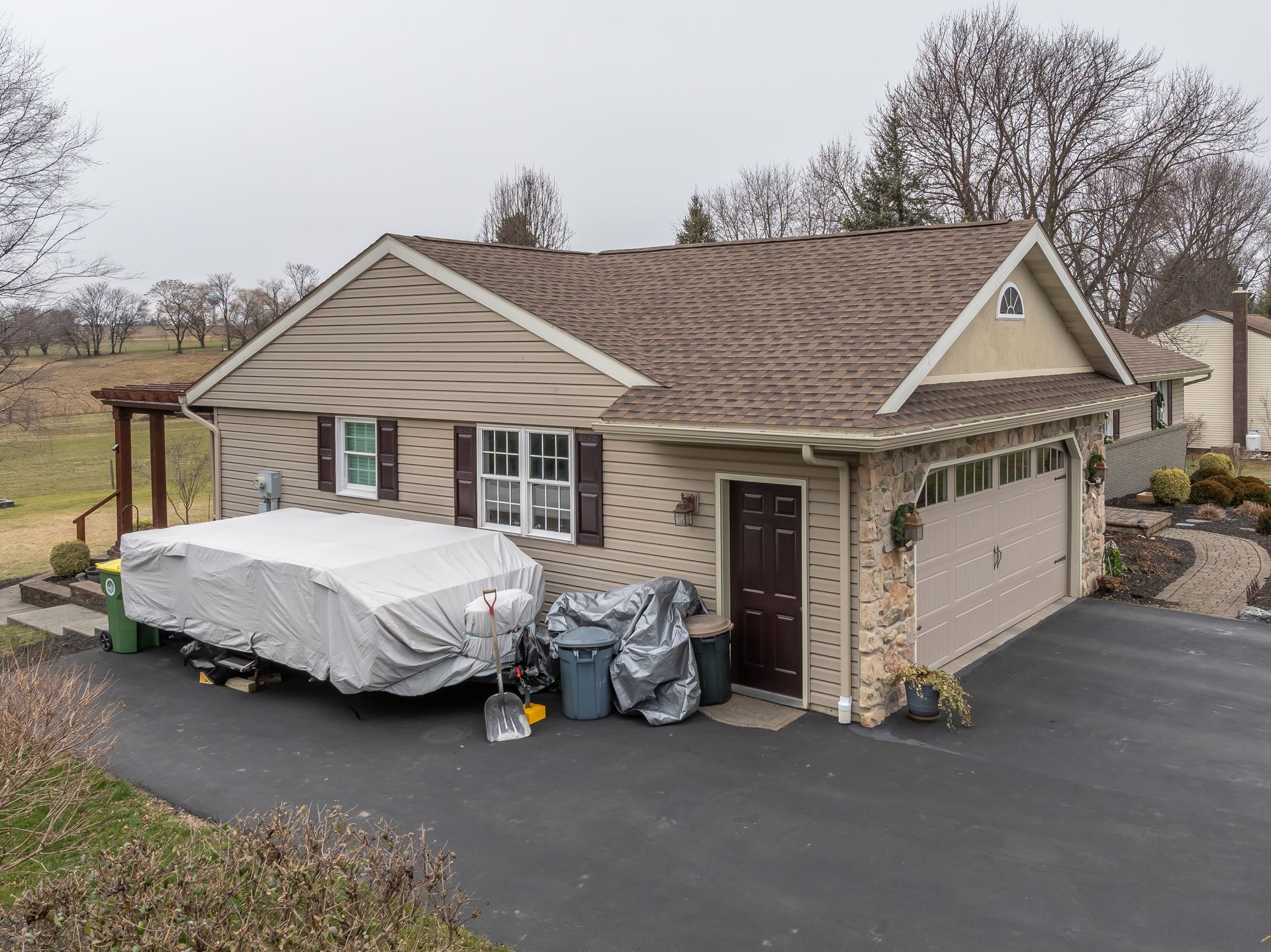 Suburban beige house with parked vehicle under tarp, trash bins, and driveway in front