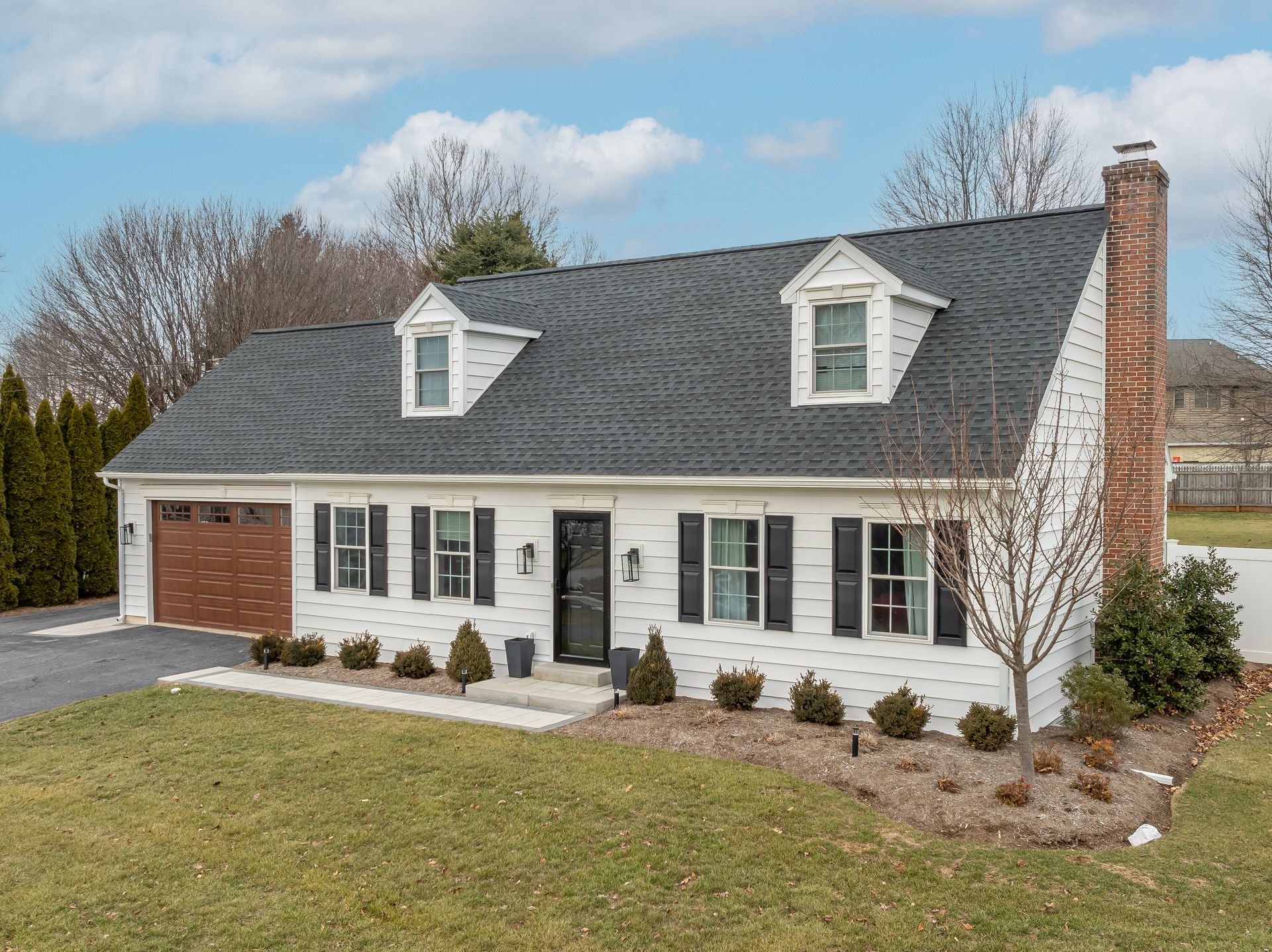 White suburban house with gray roof, brick chimney, dormer windows, and small front shrubs