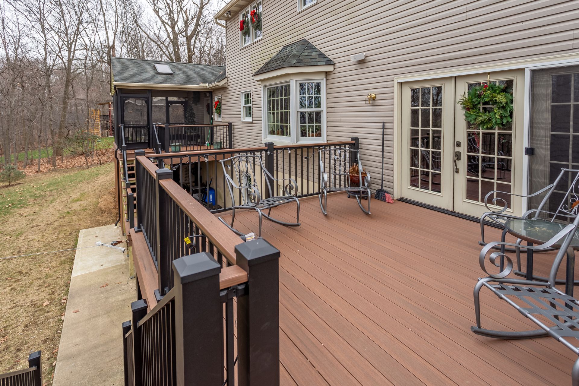 Back deck with brown railing, rocking chair, and French doors on a beige house, overlooking trees