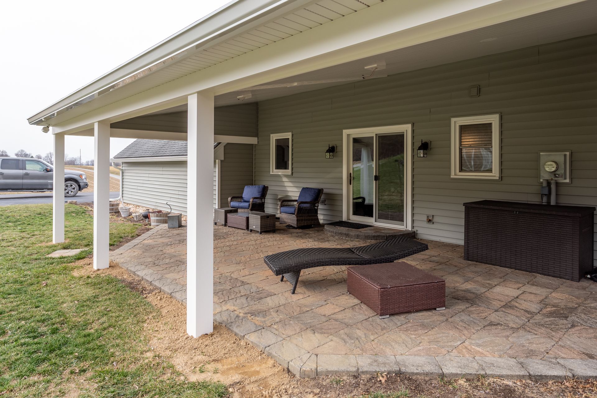 Covered patio with outdoor seating, rug, and storage chest beside a house.
