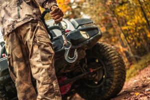 Person attaching a winch hook to a Jeep bumper on a dirt trail.