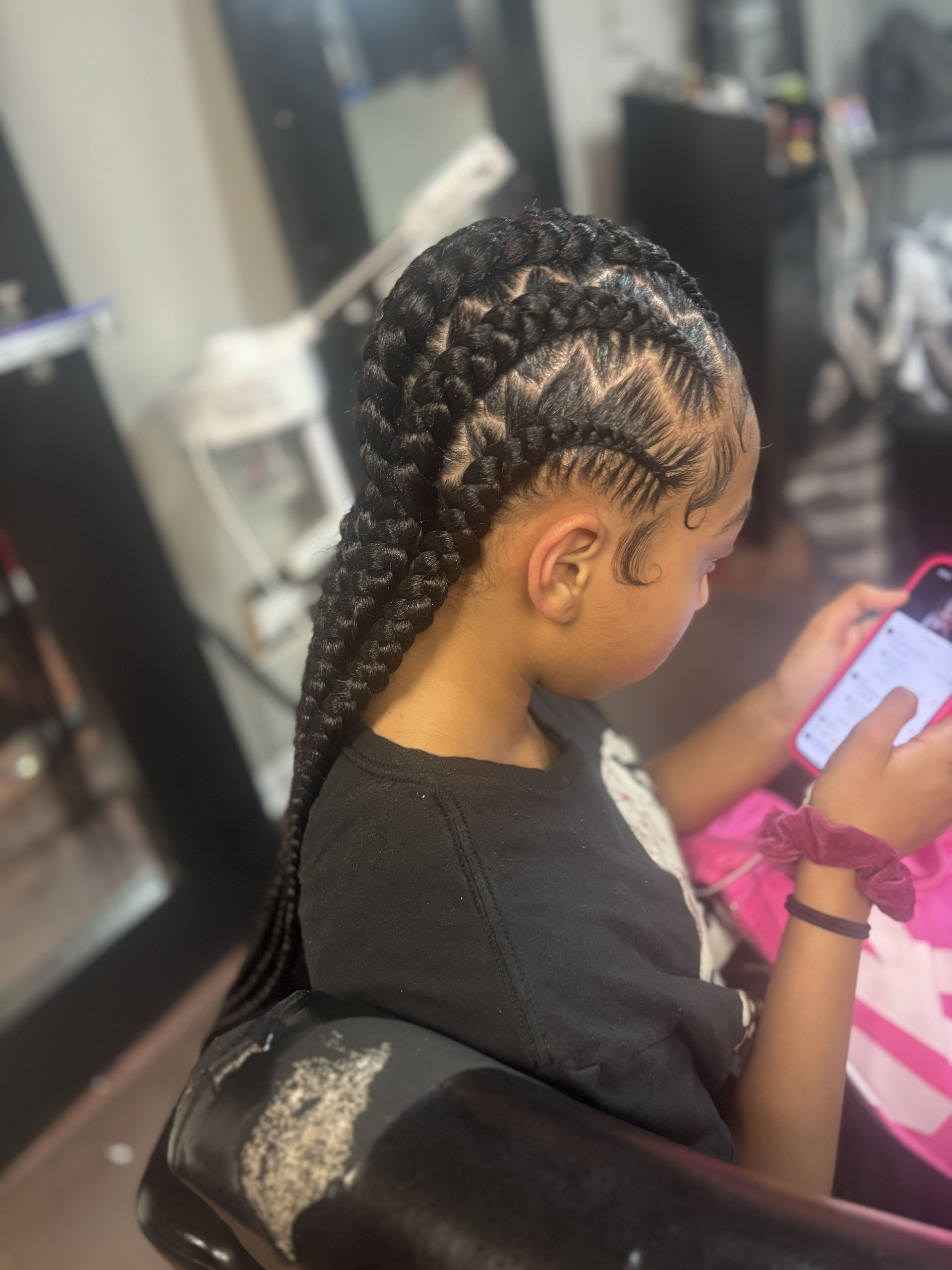 Person with braided hair sits in a salon chair, looking at a phone while getting hair styled.