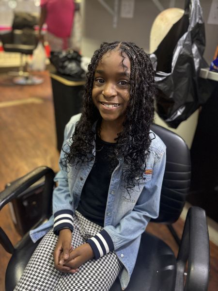 Smiling person seated in a salon chair, wearing a denim jacket over a black top and checkered pants.