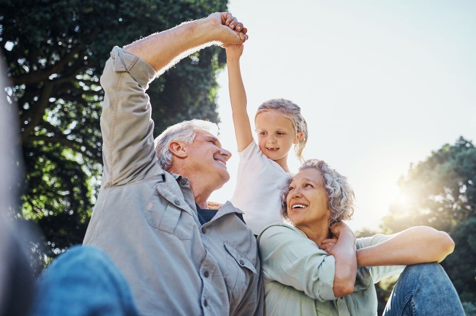 Smiling family outdoors, with a child between two adults raising joined hands in the sunlight
