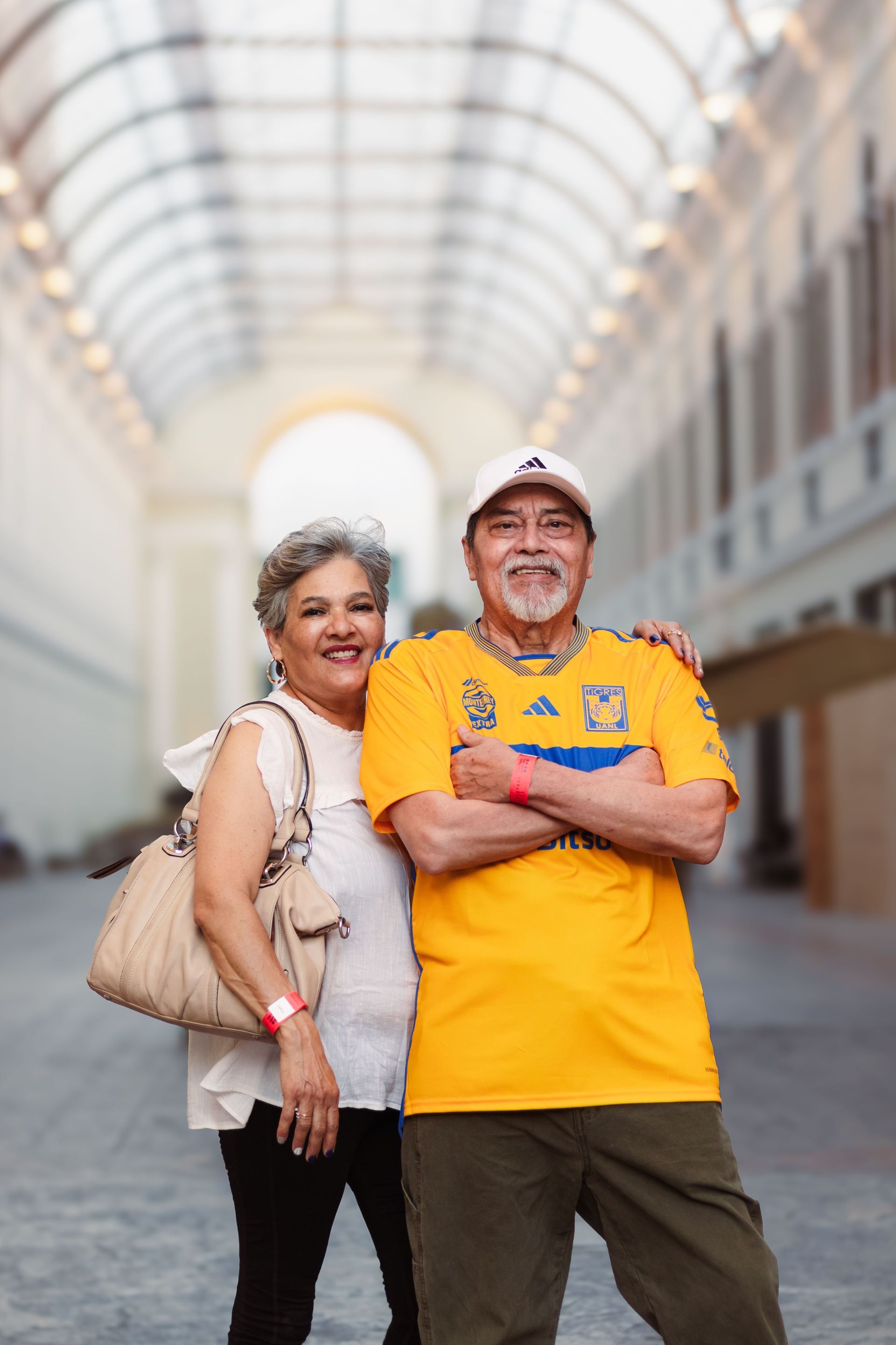 Two people smiling in a bright indoor walkway, one in a yellow shirt and cap, the other in a beige top.