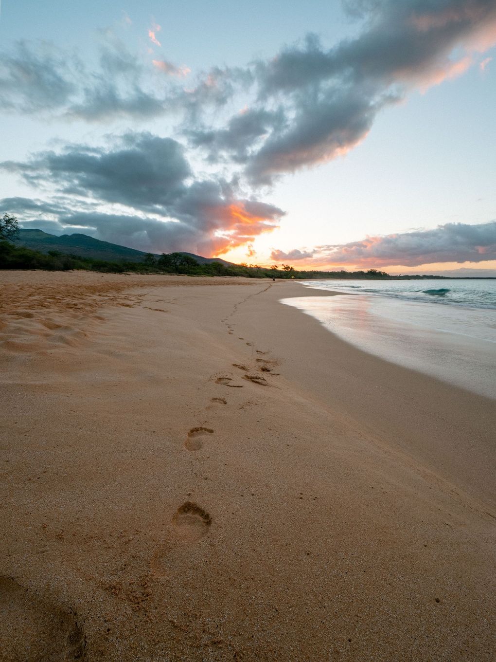 Sunset over a sandy beach with footprints, calm waves, and dramatic clouds