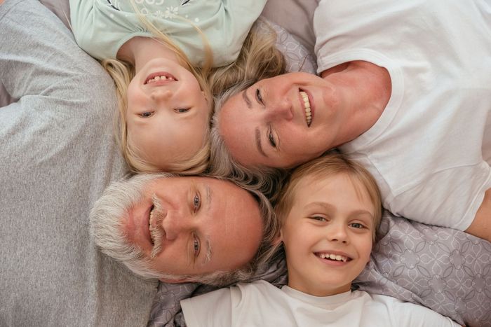 Four smiling people lying head-to-head in a circle on a bed, wearing light gray and white tops.