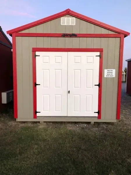 Small tan shed with red trim and white double doors on a grassy lot
