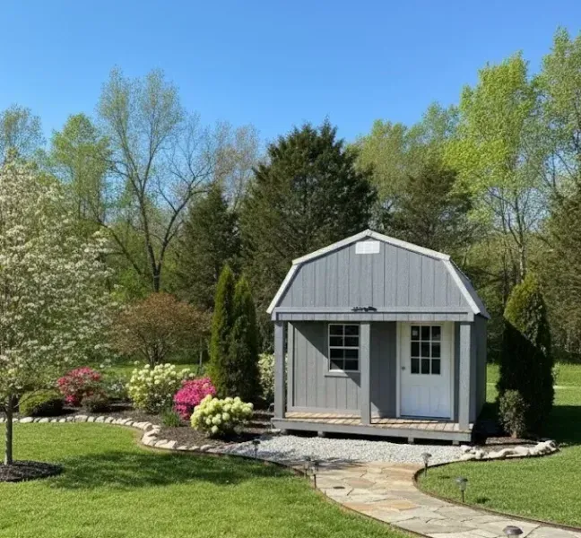 Small gray garden shed with porch, surrounded by flowers and a green lawn under a clear blue sky