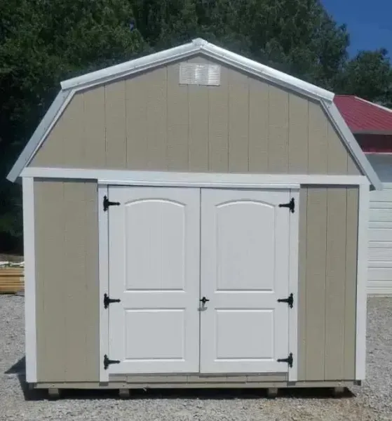 Small tan shed with white double doors and trim, standing outdoors on a sunny day.