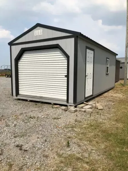 Gray prefab shed with a white roll-up door on a gravel lot under a cloudy sky
